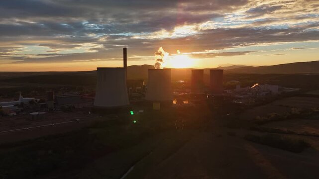 Aerial view of cooling towers and a chimney at a thermal power plant in Visonta, Hungary, releasing steam over farmland beneath a cloudy summer sunset.