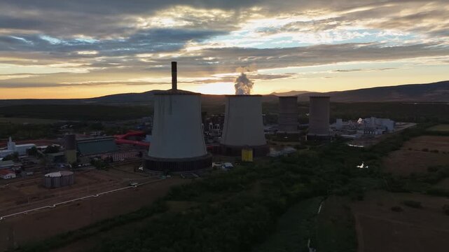 Aerial view of a lignite-fired power station with cooling towers and a chimney emitting steam at sunset, surrounded by rural fields and hills in Visonta, Hungary.