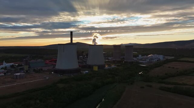 Drone view of cooling towers and a chimney at a thermal power plant releasing steam over farmland near Visonta, Hungary, beneath a cloudy summer sunset sky.