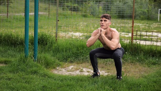 Male shirtless athlete doing squats at sports ground. Young sportsman working at summer nature. Strong sporty man exercising outdoor. Concept of active and sportive lifestyle. Slow motion
