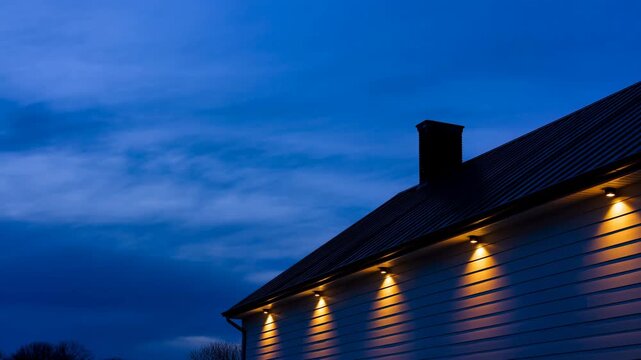 House exterior remains illuminated as clouds shift and twilight deepens above dark metal roof and brick chimney, with warm eave lights casting golden pools across horizontal siding