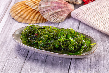 Ceramic dish holding green wakame seaweed salad alongside decorative seashells and textured cloth © Andrei Starostin