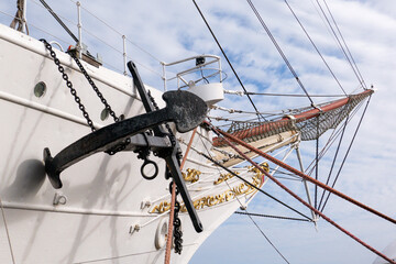 Low angle view of a historic white sailing ship bow featuring a massive black vintage anchor, rigging ropes, and golden ornamental carvings. © MarijaBazarova