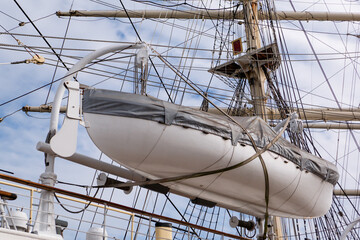 Low angle view of a historic sailing ship deck featuring a white lifeboat on davits near complex mast rigging ropes © MarijaBazarova