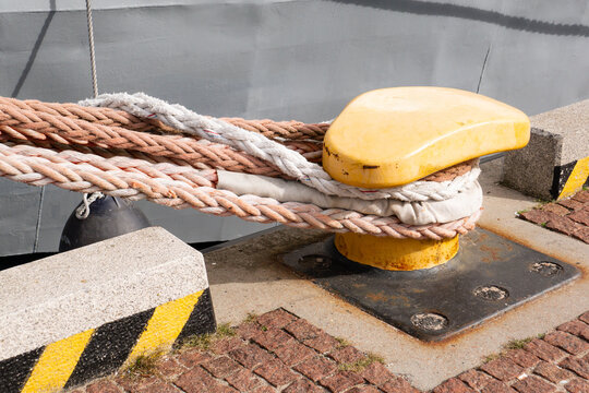 Detailed close-up shot of a heavy yellow steel mooring bollard with thick braided ropes securing a ship at a harbor pier dock.