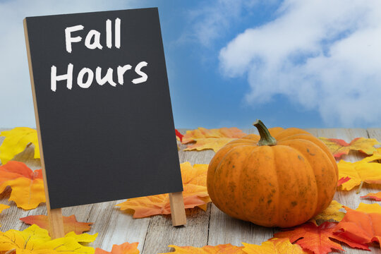 Fall Hours with fall leaves and pumpkin with black chalkboard sign on wood bench with sky