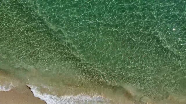 Cinematic aerial footage featuring a drone rotation and pan over crystal clear turquoise sea water, revealing parallel waves rolling from right to left over a warm brown and golden sandy beach in slow