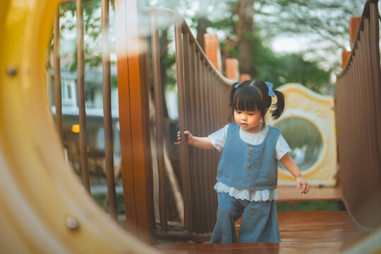 Little Asian toddler girl walking carefully on elevated playground walkway while holding safety railing for balance, exploring outdoor play equipment independently and developing coordination.
