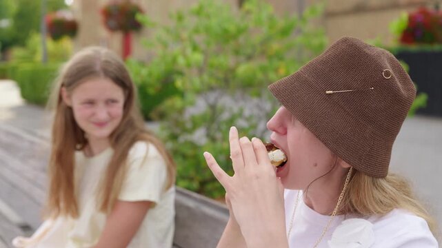 Woman eating a cream filled pastry while sitting beside a girl on a bench slow motion daughter watches her mother eat a cream puff