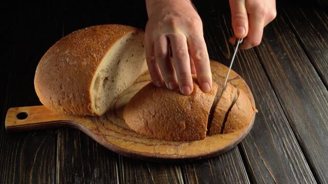 Hands slicing a round loaf of crusty bread with a knife on a wooden cutting board, creating even pieces on a dark rustic tabletop