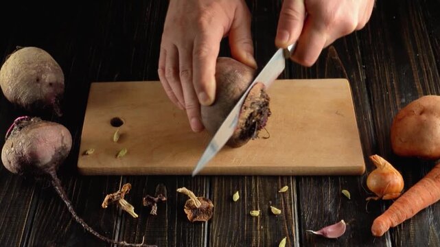 Hands slicing a whole beetroot with a kitchen knife on a wooden cutting board surrounded by vegetables and scraps in a rustic overhead food preparation scene