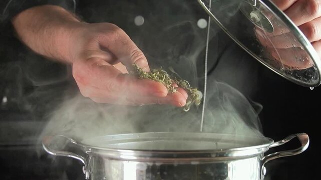 Hands adding dried herbs to a steaming stainless steel pot while lifting the lid, showing a cooking preparation process in a dark kitchen setting