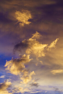 Surreal evening blue sky with white clouds with yellow glow dancing in the sky as the sun sets. In vertical format.