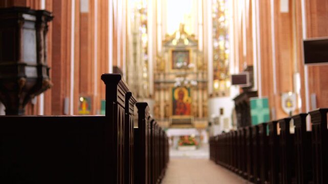 Empty church nave leads toward ornate altar, with dark wooden pews lining central aisle. Warm light brightens tall columns and gilded details, creating quiet, reverent Catholic interior.