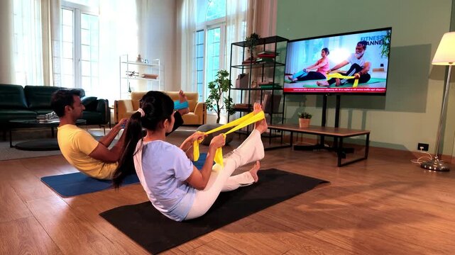 Young Indian couple sitting on the floor and stretching their legs with resistance bands held in both hands while following a TV fitness tutorial in a lavish modern home.