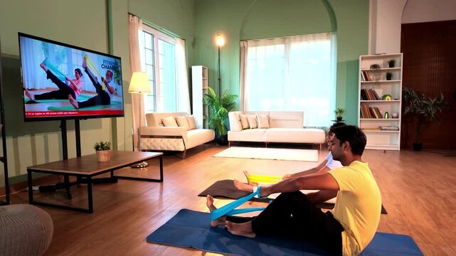 Young Indian couple sitting on the floor and stretching their legs with resistance bands held in both hands while following a TV fitness tutorial in a lavish modern home.