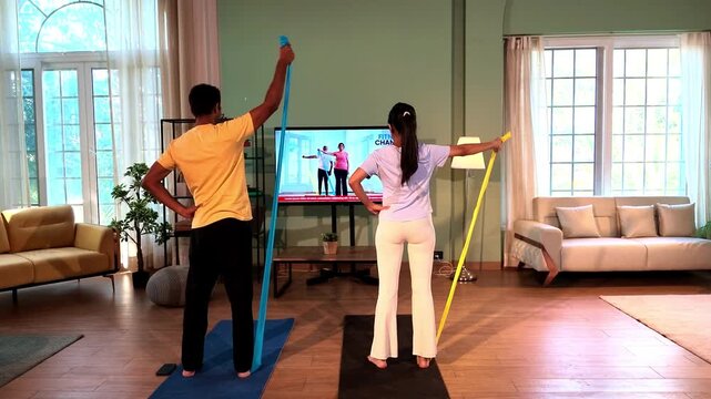 Young Indian couple performing standing shoulder raises with resistance bands anchored under their toes while following a TV fitness tutorial in a lavish modern home.