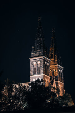 Vertical night shot focusing on illuminated twin neo-Gothic spires of Basilica of Saints Peter and Paul at Vysehrad rising above dark tree silhouettes.