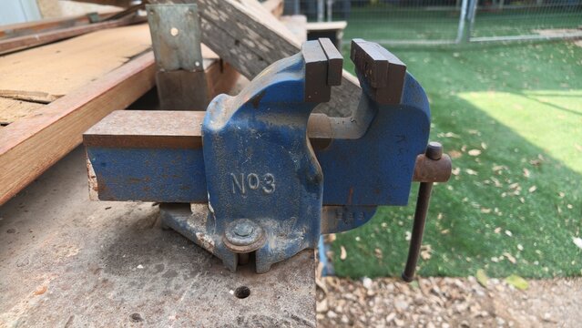 Old blue metal bench vise with number 3 mounted on a wooden outdoor workbench with planks, green grass lawn and fence blurred in the background, carpentry and woodworking workshop tool concept