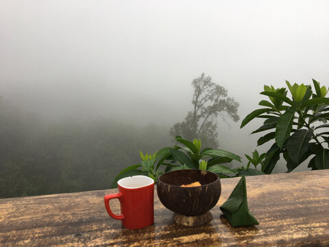 Hot coffee mug, coconut shell bowl, and traditional Thai dessert wrapped in banana leaf on a rustic wooden table with a foggy mountain background