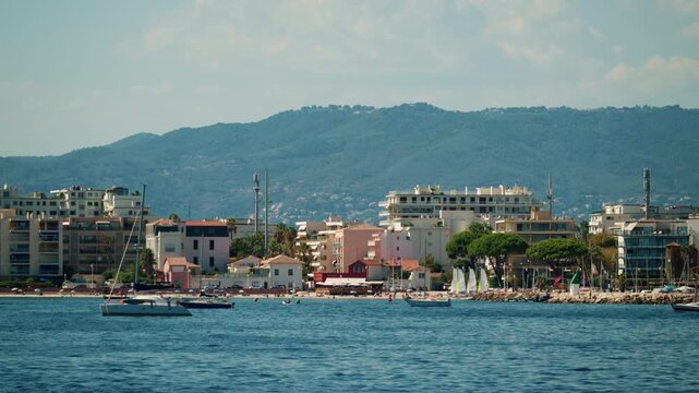 Coastal town waterfront with apartment blocks, pines and palms, sailing dinghies and an anchored sailboat, French Riviera, France.