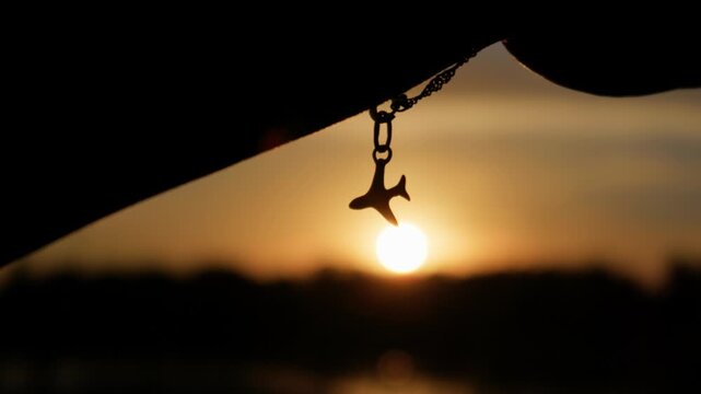 An airplane charm bracelet dangles from a wrist against a glowing golden hour sunset. Warm backlight creates a silhouette with soft depth of field and vibrant evening ambiance.