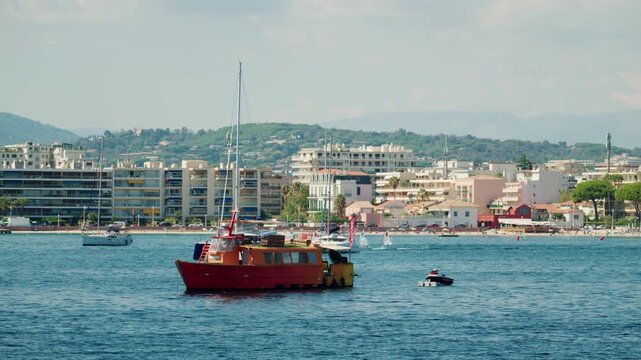 Red wooden boat anchored with a jet ski, a catamaran, sailing dinghies and a coastal town below green hills, French Riviera, France.