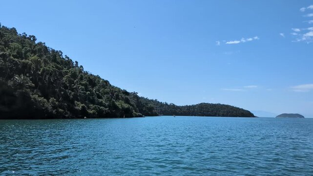 Handheld slow motion from a stationary boat of calm blue sea water in the bay of Paraty, Rio de Janeiro, with lush green tropical forested mountains stretching along the coast under a bright blue sky.