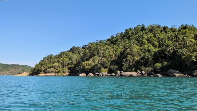 Handheld slow motion from a boat moving right showing a rocky tropical shoreline with dense green jungle and palm trees next to turquoise sea water in Paraty, Rio de Janeiro, on a sunny day.