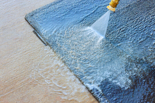 Pressure washer nozzle sprays water over dark car carpet surface outdoors, rinsing dirt toward edge