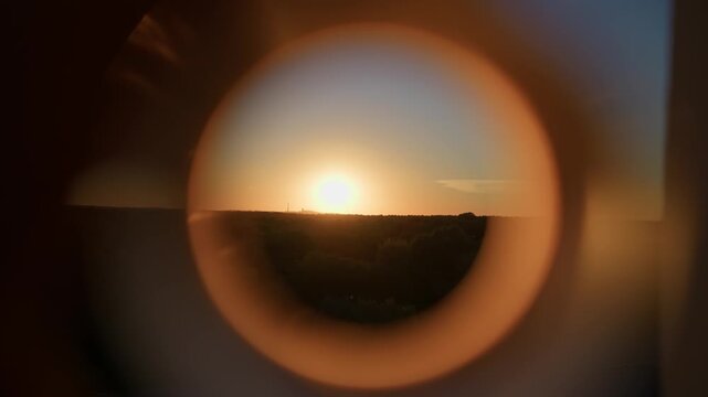 Golden sun setting over the distant forest horizon is framed by a circular opening. Warm ambient twilight creates a conceptual perspective highlighting the serene natural landscape.