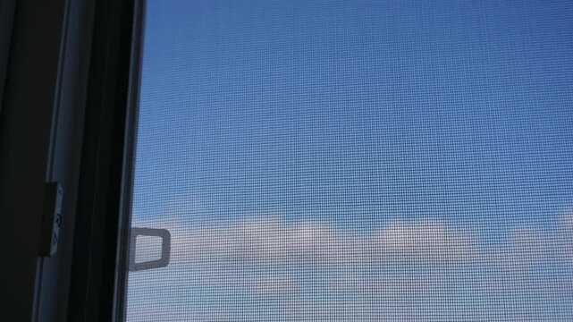 Window screen netting against blue sky provides domestic insect protection. The fine mesh texture offers ventilation while panning across the bright residential apartment window.