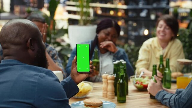 Vertical green screen smartphone mockup is held above a rooftop dining table beside a burger on a plate, offering editable screen replacement while friends toast and socialize behind the phone.