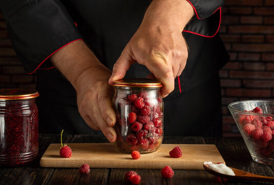 Cook fastens lid on jar of fresh raspberries, holding glass container steady on cutting board. Berry preserving takes place at dark kitchen counter with bowl of fruit nearby, closing jar for storage.