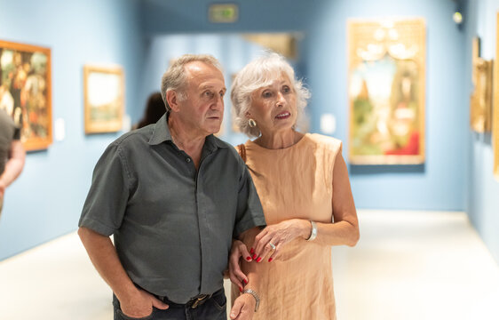 Impressed senior couple observing artwork in gallery with classic pictures visible in background