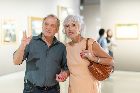Impressed senior couple observing artwork in gallery with classic pictures visible in background