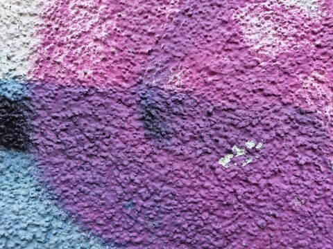 A richly bumpy paint surface blends magenta and violet, with light blue and black spray at the left edge. A small chip of cream paint has flaked away near the center.