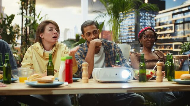 Friends gesture and debate while watching a sports match on a video projector at a rooftop barbecue, sharing suspense and spectator reactions during an outdoor party with food and drinks.