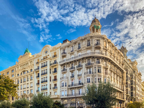 Elegant historic apartment buildings featuring classical architecture, ornate balconies, and a prominent corner dome under a blue sky with white clouds in Plaza del Ayuntamiento, Valencia, Spain.