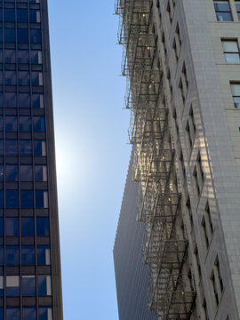 CHICAGO, IL, JUN 19, 2026: Looking up between a dark glass office tower and a stone building draped with metal fire escapes, with a distant ribbed skyscraper and sun glow in a blue sky