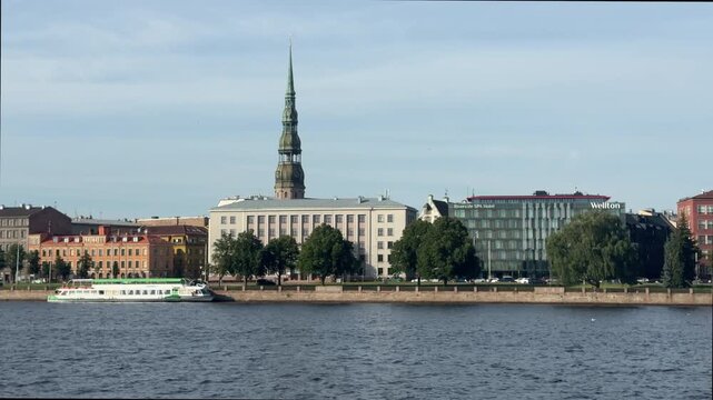 Tracking shot along the Daugava River in Riga, Latvia, capturing the historic St. Peter's Church spire, river cruise boat, and city waterfront.
