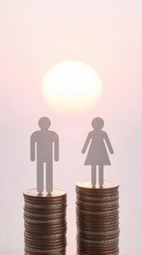Male, female paper figures stand on different height coin stacks at sunrise. Concept of gender wage gap, financial inequality in business. Man stands on higher money pile than woman, showing social