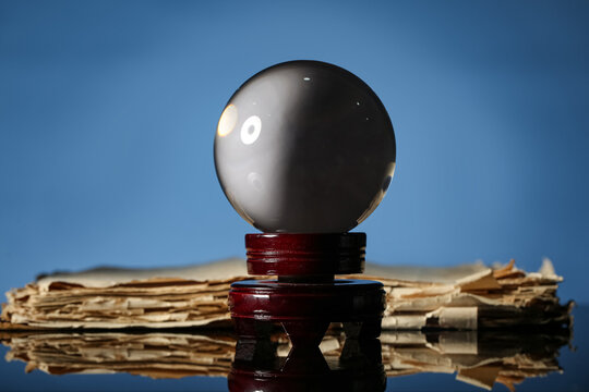 Future prediction and fortune telling concept, crystal ball on wooden stand with antique weathered book on reflective surface against blue background