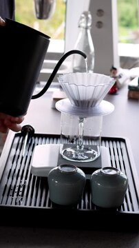 Vertical close-up of pouring hot water from a gooseneck kettle into a coffee dripper with steam rising and ceramic cups in the foreground