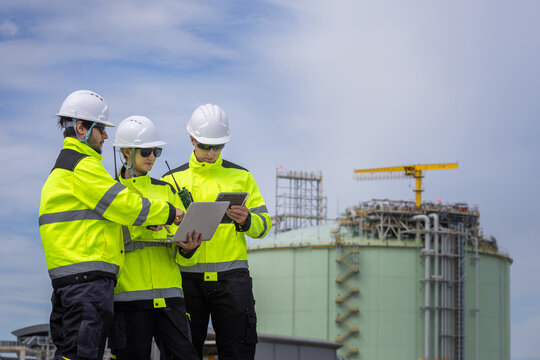 Engineers in safety jackets discussing project data with laptop outside gas storage tank plant, Team of industrial inspectors holding laptop and tablet near large storage tank outdoors