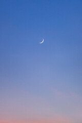 A slender crescent moon glows in a deep blue twilight sky above a softly colored horizon © narongcp