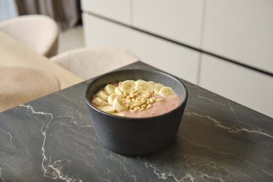 Bright and inviting banana-strawberry smoothie sits in a dark bowl, garnished with fresh banana slices and pine nuts. The cozy kitchen provides a warm backdrop for this healthy treat