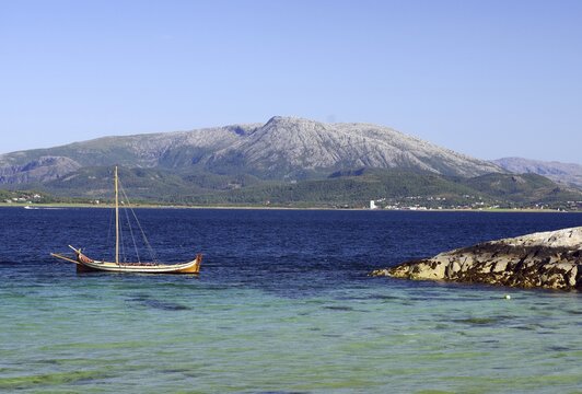 A wooden boat floats on a clear blue sea in front of a large mountain, Bronn&ouml;ysund, Nordland, Rv 17, Kystriksveien, Norway
