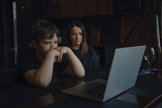 A boy sits focused at a table, watching a laptop screen closely while his mother looks on with a supportive expression