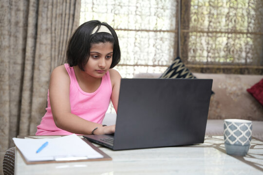 A young Indian schoolgirl using a laptop to search for information and complete her school homework at home. A focused child studying online, preparing school assignments, writing notes in a notebook.